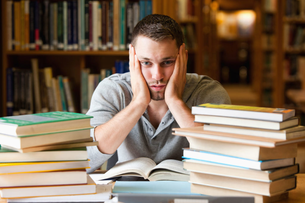 Junger Mann mit vielen Büchern vor sitzt in der Bibliothek und stützt die Hände in den Kopf